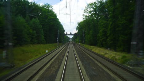 train-track-surrounded-by-lush-green-trees-and-grass-tracks-are-lined-with-overhead-power-lines-moving-forward-view-from-the-back-in-far-another-train-is-coming-forward-traveling-to-a-different-city