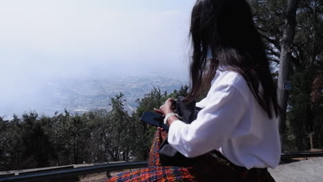 Raven-haired-woman-in-pleated-skirt-sits-at-viewpoint-above-foggy-city