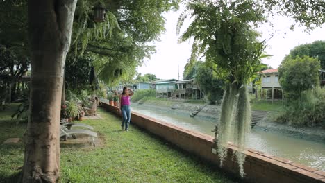 Thai-Lady-Walking-in-a-Peaceful-Garden-Next-to-a-Canal-in-Ayutthaya,-Thailand