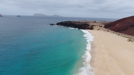 La-Graciosa-island:-aerial-view-traveling-in-along-the-shore-of-Las-Conchas-beach-on-a-sunny-day-and-turquoise-waters