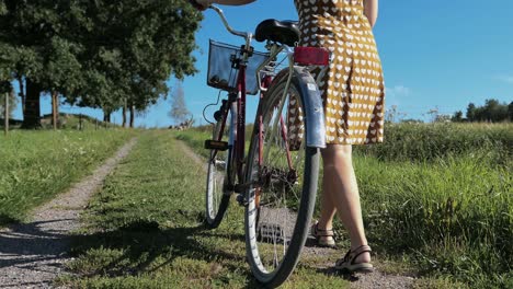 Woman-in-vintage-dress-walking-with-bike-country-road-in-countryside,-crane-shot