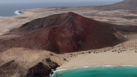 Las-Conchas-beach,-La-Graciosa-island:-aerial-view-in-orbit-to-the-Bermeja-mountain-in-Las-Conchas-beach-on-a-sunny-day-and-turquoise-waters