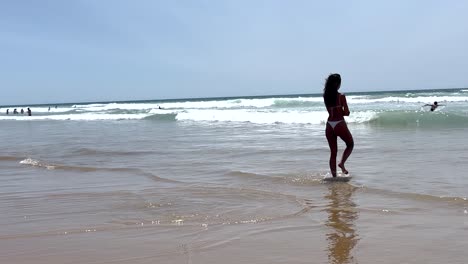 A-woman-in-a-bikini-cautiously-dips-her-feet-into-the-ocean-water,-expressing-hesitation-about-the-water's-temperature-while-enjoying-the-seaside-experience