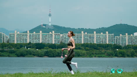 Athletic-Pretty-Woman-in-20th-Exercises-with-Cordless-Jump-Rope-Jumping-on-One-Leg-and-Both-Legs-ina-Grassy-Riverbank-Meadow-at-Han-River-Park-With-N-Seoul-Namsan-Tower-Behind---Profile-view-Slowmo
