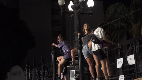 Group-of-young-women-at-night-climb-over-public-building-gates-to-take-group-pictures,-one-drinks-alcohol