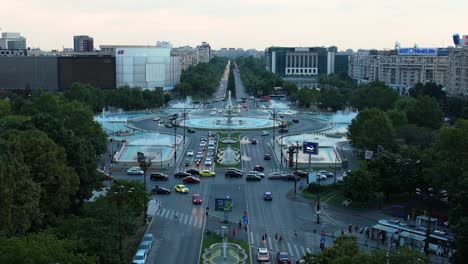 Aerial-View-Over-Unirii-Square-With-The-Famous-Water-Fountains-In-The-Main-Frame,-At-Dusk,-Bucharest,-Romania,-Heavy-Traffic,-Cars-Passing-By,-Blue-Water-Fountains