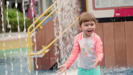 Adorable-Toddler-Girl-Reaching-Out-to-Touch-Fountain-Water-Sprayed-at-Outdoor-Playground
