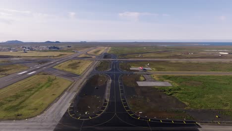 Iceland-international-airport-with-distant-airplane-taking-off-on-sunny-day,-aerial