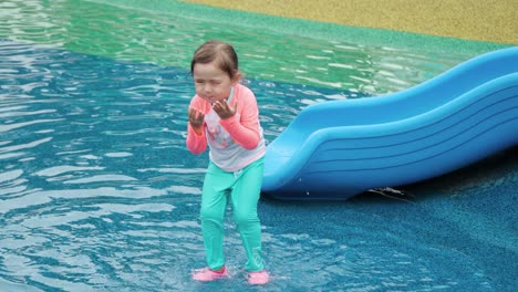 Cute-Toddler-Girl-in-Aquapark-Jumps-in-Shallow-Puddle-Pool-Happily-Like-a-Frog,-Splashes-Water-in-Eyes-and-Wipes-Face-With-Hands-Playing-At-Water-Park-Playground