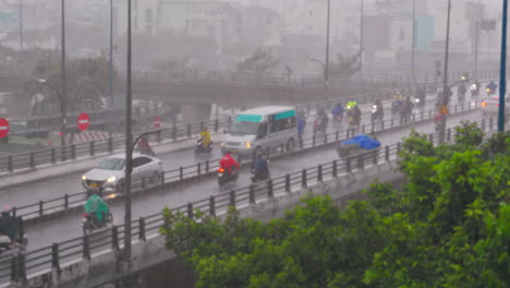 Heavy-rain-traffic-on-city-bridge,-vehicles-passing-with-buildings-and-trees-in-the-background