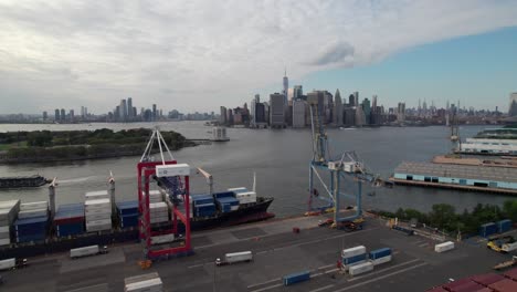 Large-container-ship-being-loaded-with-Manhattan-skyline-in-the-background,-4K-aerial