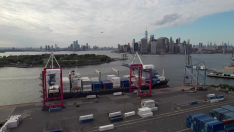 Container-ships-being-loaded-in-New-York-harbor,-industrial-scene-with-Manhattan-skyline-in-distance,-4K-aerial