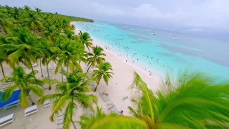 FPV-flight-over-palm-trees-and-sandy-beach-with-many-tourist-Swimming-in-Caribbean-sea-during-sunny-day,-Saona-Island