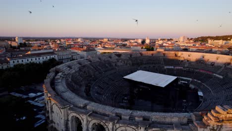 drone-aerial-shot-of-the-famous-arena-in-Nimes,-a-city-in-the-south-of-france,-front-and-interior-view-with-the-sunset-off-camera-illuminating-the-city,-quiet-streets-and-a-few-trees-and-birds