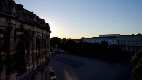 aerial-drone-shot-of-the-famous-arena-in-Nimes,-a-city-in-the-south-of-France,-seen-from-the-side-with-the-sunrise-on-the-horizon-and-a-clear-sky,-quiet-streets-and-a-few-trees
