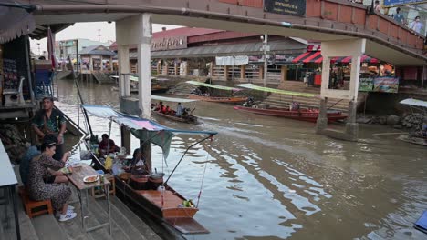 The-camera-pans-from-right-to-left-to-a-group-of-tourists-eating-their-dinner-before-they-board-the-board-for-their-night-tour-of-Amphawa-Floating-Market-in-Samut-Songkhram,-Thailand