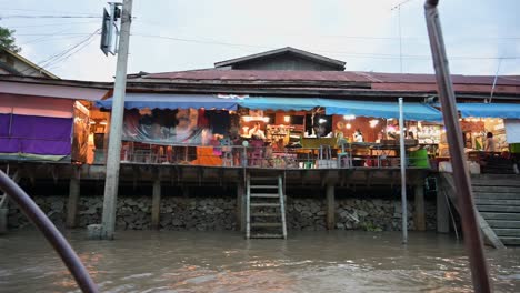 A-wooden-boat-passing-by-rows-of-restaurants-and-souvenir-shops-by-the-canals-of-a-popular-tourist-destination,-the-Amphawa-Floating-Market-in-Samut-Songkhram,-Thailand