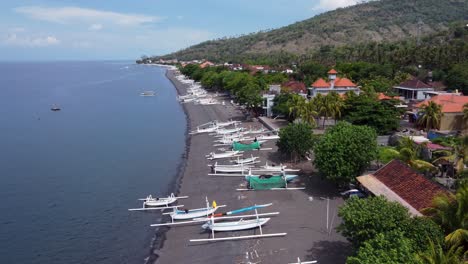 Aerial-view-of-amed-village-seaside-with-local-white-Jukung-fishing-boats-on-black-volcanic-sand-beach,-villager-houses-and-mountains-in-the-background