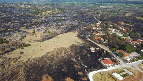 Aerial-of-village-surrounded-by-burnt-land-in-the-aftermath-of-a-wildfire-in-northern-Greece,-August-2023