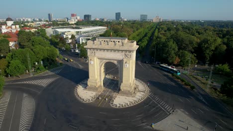 Cinematic-Rotating-View-Of-The-Arch-Of-Triumph-With-The-House-Of-The-Free-Press-In-The-Distance,-Surrounded-By-Lush-Green-Vegetation-And-Tall-Office-Buildings,-Bucharest-Romania