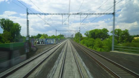 pov-fast-moving-train-with-three-tracks-view-traveling-to-a-different-stations-with-electrical-cables-above-graffiti-walls-on-the-sides-with-trees-forests-fields-and-partly-cloudy-freedom-travel