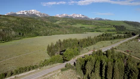 SUV-Blanco-Conduciendo-Por-Una-Carretera-De-Montaña-Entre-árboles-Durante-El-Día-Con-Montañas-Nevadas,-Antena
