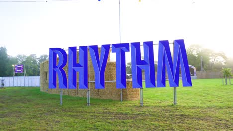 huge-RHYTHM-text-sign-of-blue-colour-realistic-3D-with-inward-lines-placed-hanging-on-metal-fence-in-the-nature-on-grassy-field-sunrise-clear-sky-clouds-plenty-bales-on-top-of-each-other-festival-area