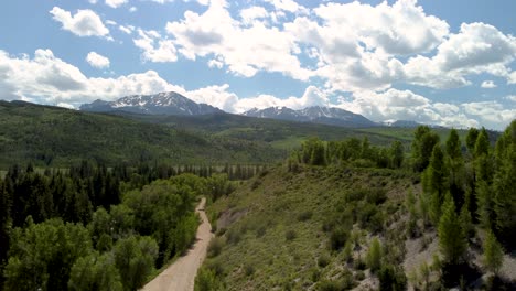 Empty-dirt-road-leading-to-the-mountains-with-pine-trees-and-a-river,-aerial