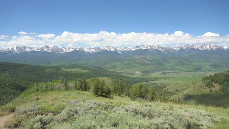 View-from-the-top-of-a-Colorado-hiking-trail-overlooking-a-green-valley-with-snowcapped-mountains,-static