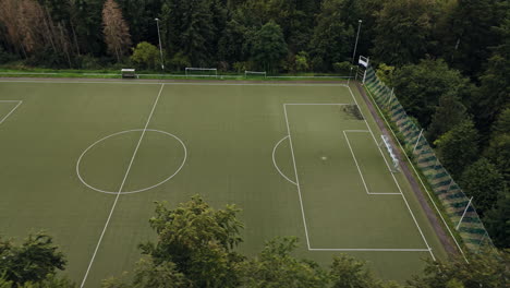 Aerial-Flight-besides-an-empty,-green-football-field-in-the-middle-of-the-forest,-Drone-Footage