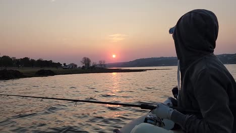 A-woman-fishing-from-a-small-boat-on-a-lake-with-the-sun-setting-in-the-distance