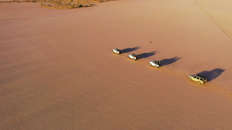 Drone-shot-tracking-a-queue-of-4x4-vehicles,-driving-across-distant-desert-in-sunny-Saudi-Arabia