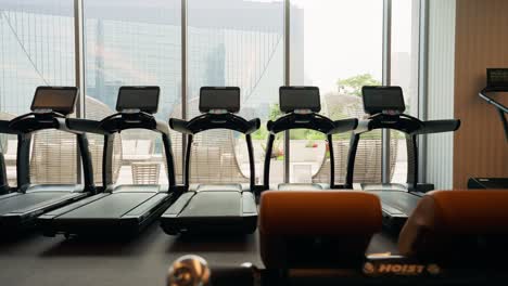 Panning-across-multiple-treadmills-in-a-fitness-center,-situated-in-front-of-expansive-windows-that-provide-a-view-to-the-outdoors