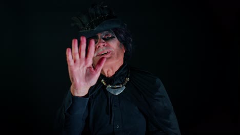 Frightening-senior-man-with-Halloween-witcher-makeup-waves-hand-palm-in-hello-hi-gesture-welcomes