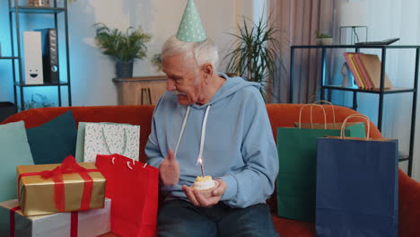 Happy-senior-old-man-celebrating-birthday-party,-makes-wish-blowing-burning-candle-on-small-cupcake