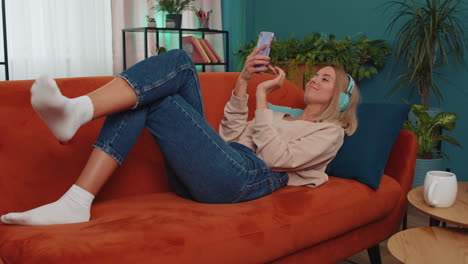 Happy-young-woman-in-wireless-headphones-relaxing-lying-on-sofa-at-home-listening-favorite-music