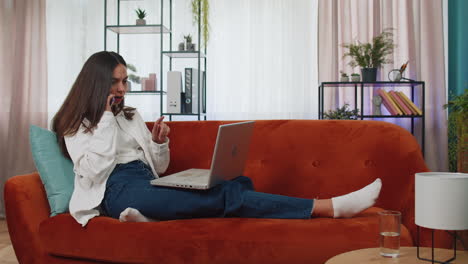 Young-woman-using-laptop-computer-sitting-on-sofa-working,-online-shopping-from-home-office