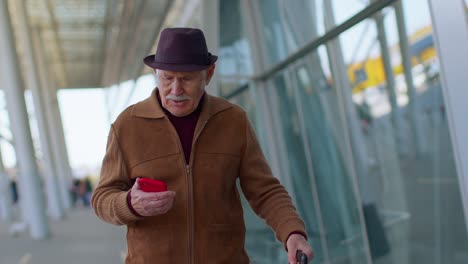 Abuelo-Turista-Senior-Hombre-Caminando-En-El-Vestíbulo-Del-Aeropuerto-Internacional,-Usando-Teléfono-Móvil,-Enviando-Mensajes-De-Texto