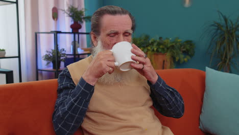 Attractive-smiling-senior-old-grandfather-man-drinking-a-cup-of-coffee-or-herbal-tea-at-home-couch