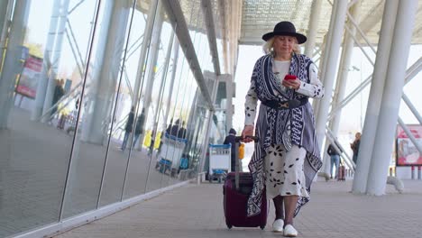 Senior-tourist-grandmother-woman-walking-on-international-airport-hall,-using-mobile-phone,-texting
