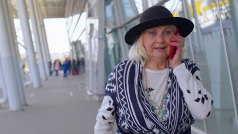 Mujer-Abuela-Turista-Senior-Caminando-Por-El-Hall-Del-Aeropuerto-Internacional,-Usando-Teléfono-Móvil,-Hablando