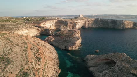 Drone-is-flying-over-clear-turquoise-waters-of-Malta's-Blue-Lagoon,-unveiling-rugged-rock-formations-and-coastal-caves-along-Como-Island's-captivating-shoreline
