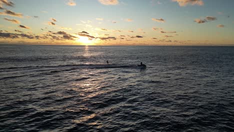 Drone-shot-showing-silhouette-of-cruising-jet-ski-carrying-surfer-on-ocean-water-during-golden-sunset-time---Wild-Bird-flying-into-camera