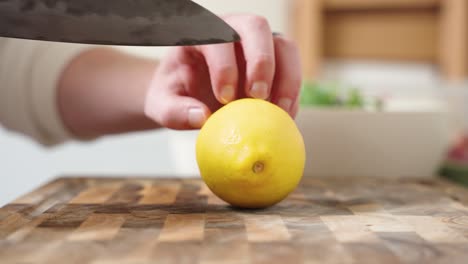 A-Male-Hand-Holding-a-Knife-Cuts-is-cutting-a-Fresh-Organic-Lemon-into-in-a-Cross-Section-Half-on-a-Wooden-Cutting-Board-inside-a-White-Minimalist-Kitchen