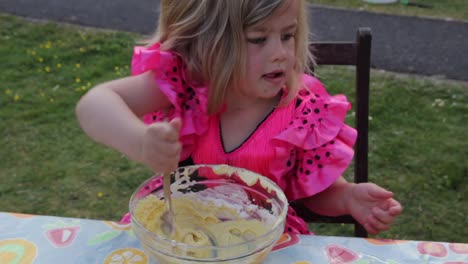 Mid-shot-of-enthusiastic-young-girl-in-pink-princess-dress-mixing-a-bowl-of-cake-mix-on-a-patio-table-in-daytime
