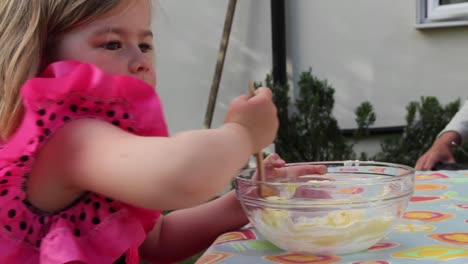 Mid-shot-of-young-girl-in-a-pink-princess-dress,-stirring-a-bowl-of-cake-mix-as-a-parent-adds-a-spoonful-of-sugar