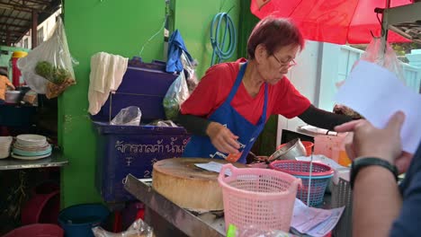 Eine-Straßenhändlerin-In-Bangkok-Liest-Die-Bestellungen-Ihrer-Kunden-Für-Gegrilltes-Hähnchen-Und-Schweinefleisch,-Das-Sie-Zubereiten-Muss