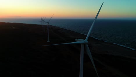 Wind-turbine-line-at-blue-hour-dusk-by-ocean-side,-golden-hour-sunset,-aerial-drone-orbit