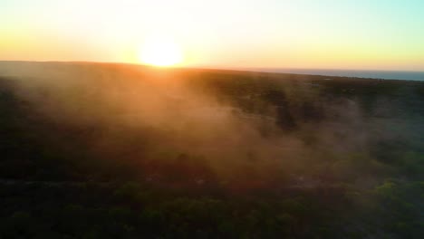 Misty-haze-fog-smoke-rises-spread-across-shrub-landscape-at-golden-hour-orange-glow-in-air