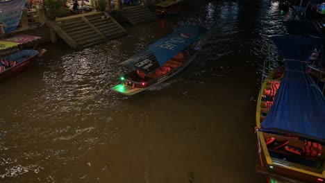 A-night-tour-on-a-boat-ride-cruising-from-the-top-right-side-of-the-frame-to-the-bottom-left-side-in-the-canal-of-Amphawa-Floating-Market-in-Samut-Songkhram,-Thailand